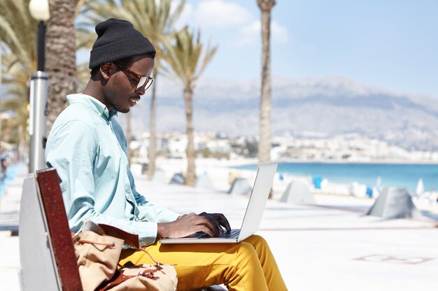 Stylish young writer in hat and shades typing on keyborad on laptop