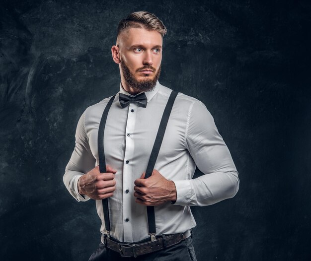 Stylishly dressed young man in shirt with bow tie and suspenders. Studio photo against a dark wall background