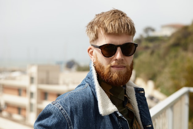Free photo stylsih unshaven young caucasian man in black rectangular sunglasses and denim jacket relaxing
