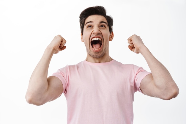 Free photo success and celebration. excited man looking up at screen, winning bet, shaking hands and scream yes, celebrating victory, win and triumphing, standing over white background