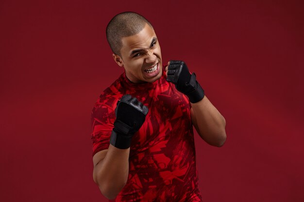 Success, determination, challenge and competition concept. Picture of furious aggressive young Afro American fighter wearing black training gloves boxing in studio, roaring, preparing for fight