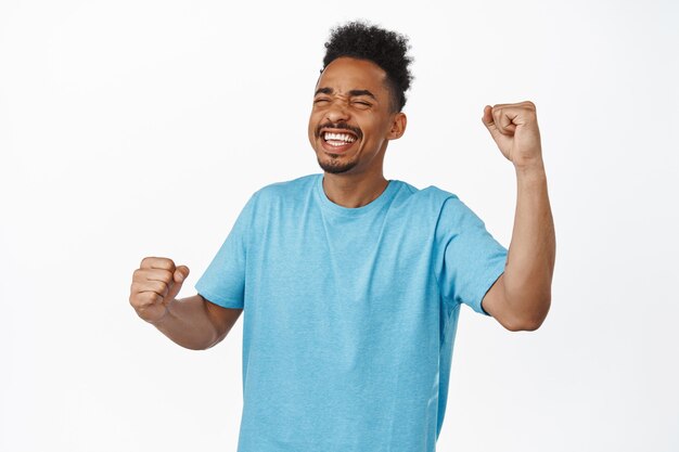 Success. Happy african american guy, sport fan chanting, smiling satisfied, achieve goal, rooting for team, pleased with good score, fist pump in triumph, standing on white.