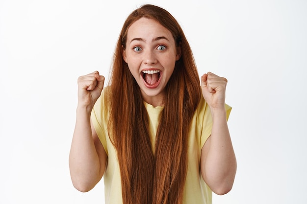 Evviva il successo. felice ragazza rossa che canta, celebra la vittoria, urla di eccitazione e gioia, salta come un vincitore e sembra sorpresa, sfondo bianco.