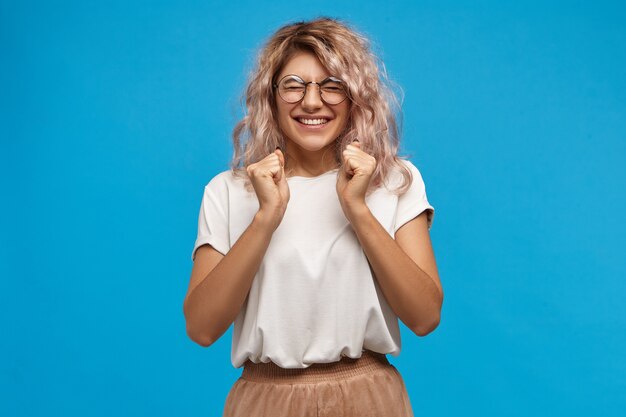 Success, positive news, joy and happiness concept. Ecstatic excited adorable stylish girl in trendy round glasses smiling broadly and clenching fists