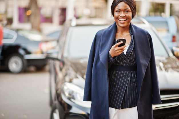 Success stylish african american woman in coat against black business suv car looking at phone