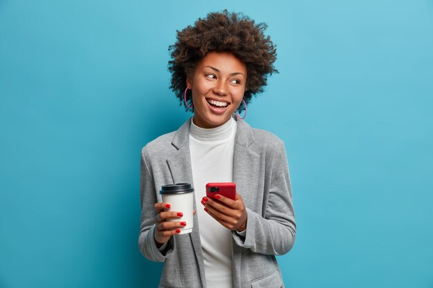 Successful afro american entrepreneur in formal wear holds mobile phone, orders lunch online, drinks takeaway coffee, checks message, makes new appointment, looks with broad shining smile aside