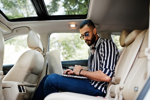줄무늬 셔츠와 선글라스를 입은 성공적인 아랍 남자는 손에 노트북을 들고 흰색 suv 차 안에서 포즈를 취합니다.