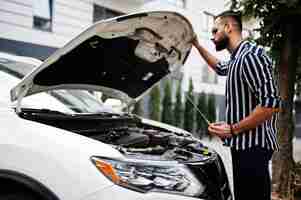 Free photo successful arab man wear in striped shirt and sunglasses pose near his white suv car check engine with open hood