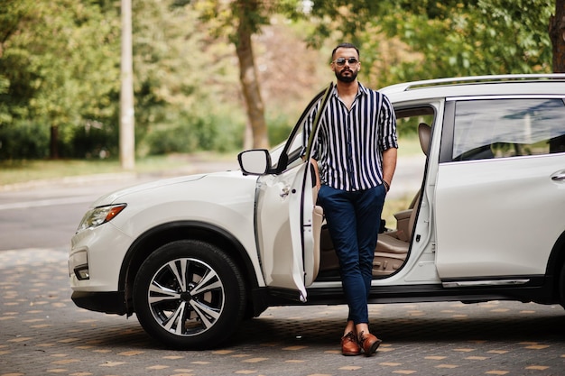 ストライプのシャツとサングラスで成功したアラブ人は彼の白いsuv車の近くでポーズをとる輸送中のスタイリッシュなアラビア人男性