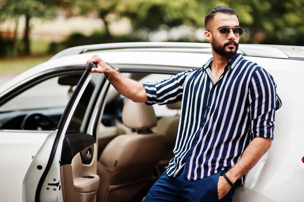 Successful arab man wear in striped shirt and sunglasses pose near his white suv car Stylish arabian men in transport