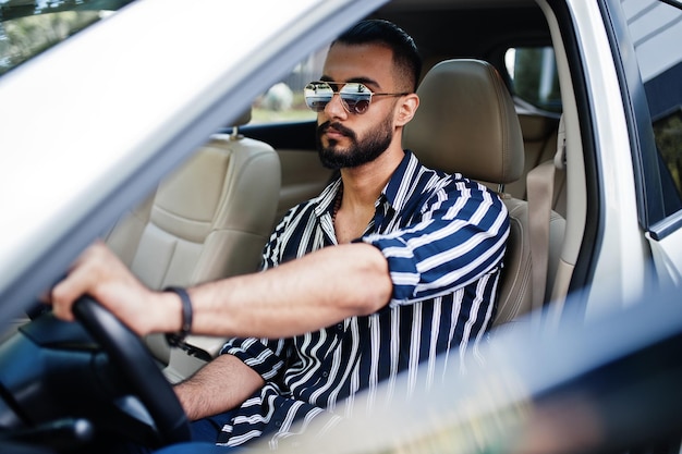 L'uomo arabo di successo indossa una camicia a righe e occhiali da sole pongono al volante della sua auto bianca suv eleganti uomini arabi in trasporto