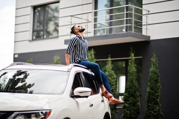 Successful arab man wear in striped shirt and sunglasses sitting on the roof of his white suv car