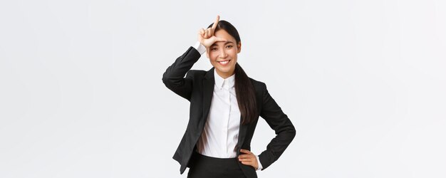 Successful asian businesswoman in black suit mocking her rivals showing loser sign as winning and celebrating victory over competitors standing pleased over white background