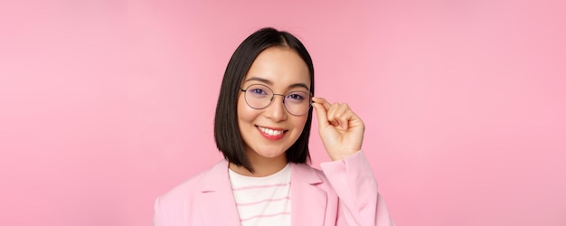 안경을 쓴 성공적인 아시아 여성 사업가가 분홍색 배경 위에 정장을 입은 카메라를 보고 웃고 있다