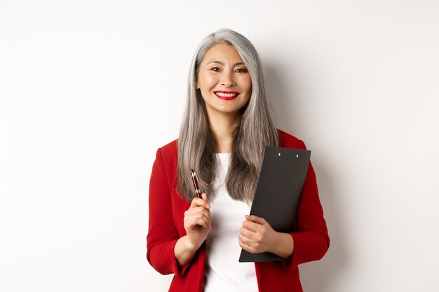 Успешный азиатский босс леди в красном пиджаке, держа доску сзажимом для бумаги с документами и ручкой, работая и выглядя счастливым, белым фоном.