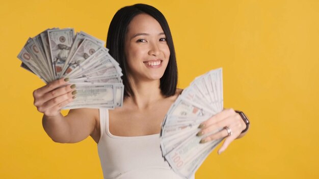 黄色の背景に分離されて幸せそうに見えるカメラでお金でポーズをとって成功したアジアの女性