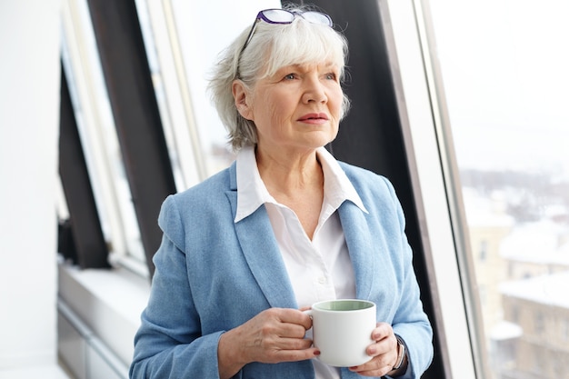 Successful attractive mature female architect wearing eyeglasses on her head and formal suit enjoying small break, drinking coffee by window, holding mug and looking with thoughtful facial expression