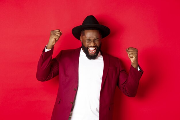 Successful Black man in suit saying yes, rejoicing of winning or achieving goal, raising hands up and screaming of joy, red background