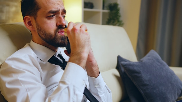 Успешный бизнесмен ест гамбургер, сидя на диване после утомительного дня и разговаривает по телефону.