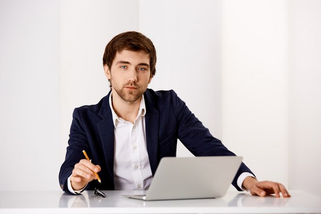 Успешный бизнесмен сидят в своем кабинете, работают над проектом с ноутбуком, держат карандаш и смотрят