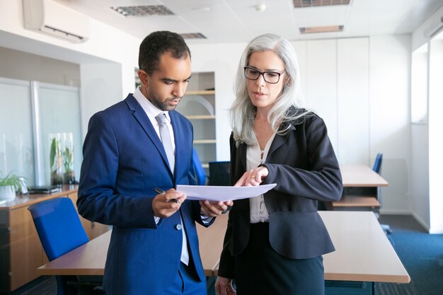 Successful businessman in suit reading document for signing and female grey-haired manager in eyeglasses pointing at something in report. Partners working in office. Business and management concept