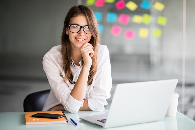 Успешная деловая женщина работает на портативном компьютере и думает над новыми идеями в своем офисе, одетая в белую одежду