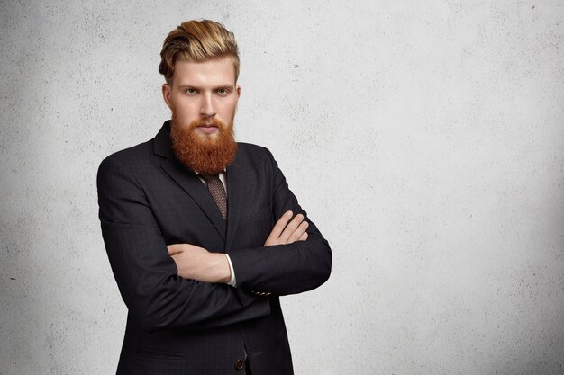 Successful and confident young entrepreneur or office worker with thick beard and stylish haircut wearing suit and standing with arms crossed against wall with copy space for your information