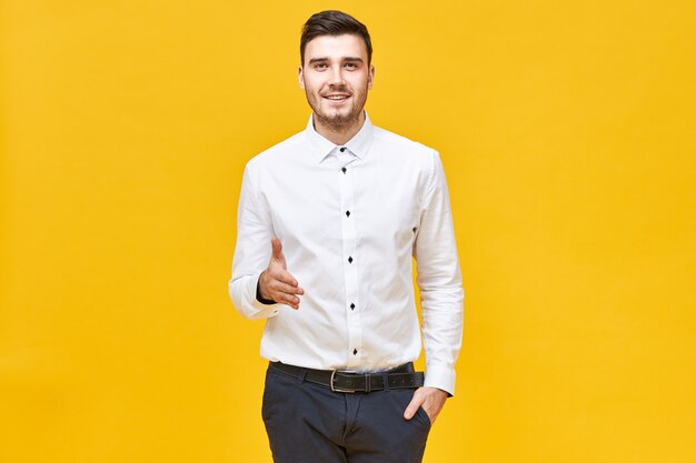 Successful confident young man in white formal shirt and classical trousers smiling and reaching out hand to shake yours, making welcoming and greeting gesture, ready to make agreement