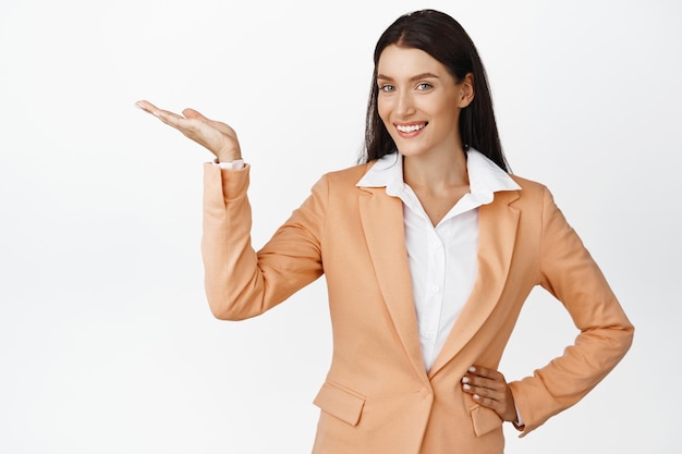 Donna aziendale di successo che dimostra il prodotto che punta allo spazio vuoto che mostra pubblicità e sorridente in piedi in tuta su sfondo bianco