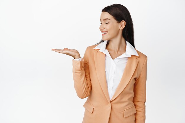 Успешная корпоративная женщина показывает объект на открытой ладони, смотрит на свою руку и довольно улыбается, стоя в костюме на белом фоне