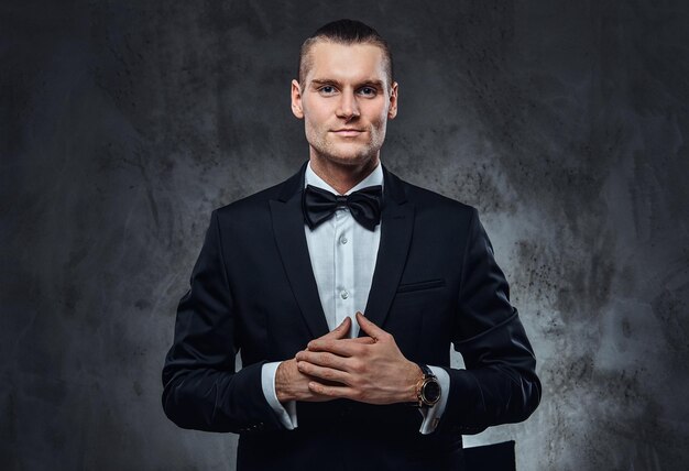 Successful elegantly dressed man wearing a black classical suit and bow tie, looking at a camera. Studio shot on a dark textured wall