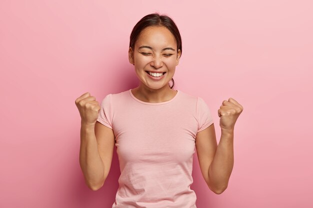 성공적인 정서적 아시아 여성은 주먹을 움켜 쥐고, 성공을 축하하고, 넓은 미소를 지으며, 눈을 감고, 흥분하고 즐겁고, 달콤한 승리의 맛을 즐기고, 캐주얼 한 옷을 입습니다.
