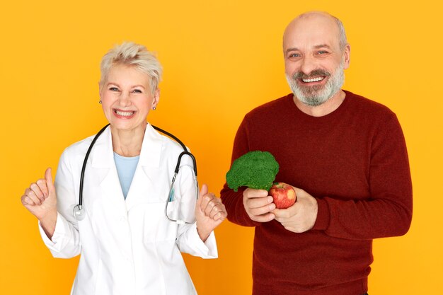 Successful energetic middle aged woman physician with stethoscope around her neck having excited facial expression, her happy senior patient choosing healthy diet, holding broccoli and apple, smiling