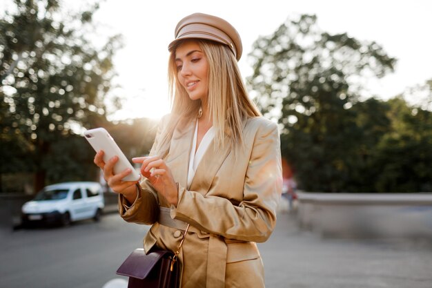 Successful fashionable european woman in elegant casual outfit posing mobyle phone outdoor. Sunset colors.