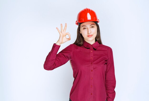 빨간색 하드 헬멧을 쓴 성공적인 여성 건축가가 서서 확인 표시를 합니다.