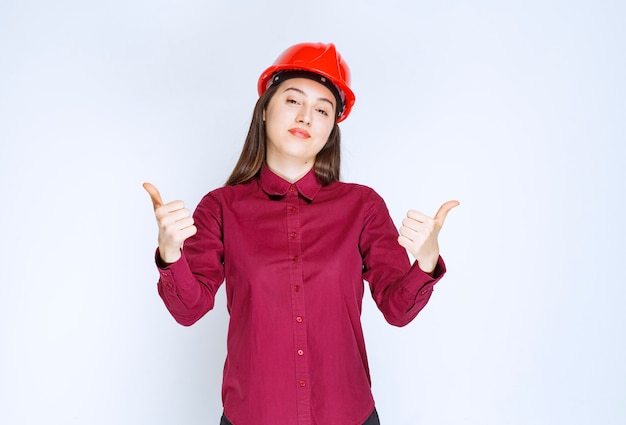 Successful female architect in red hard helmet standing and giving thumbs up. 