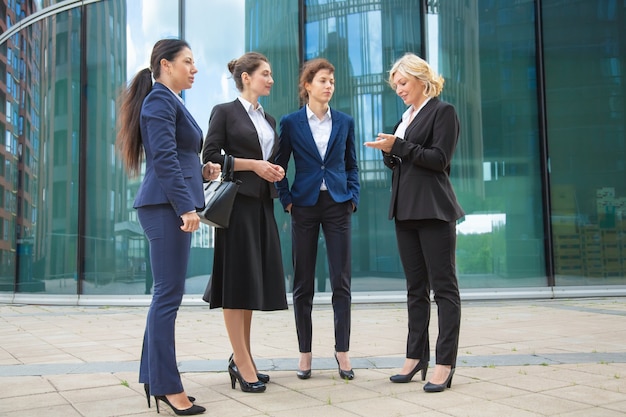 Successful female managers discussing project outdoors. Businesswomen wearing suits, standing together in city and talking. Full length, low angle. 
