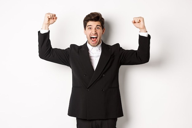 Successful handsome businessman triumphing, raising hands up and shouting yes, rejoicing over achievement, standing against white background.