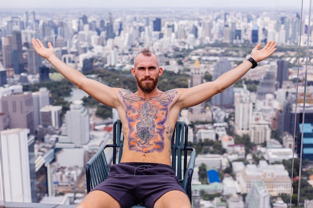 Успешный красивый европейский бородатый брутальный татуированный сильный мужчина топлес с часами сидит на стуле на высоком этаже с потрясающим видом на город