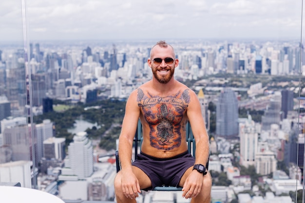 Successful handsome european bearded brutal tattooed strong man topless with watch sits on chair on high floor with amazing city view