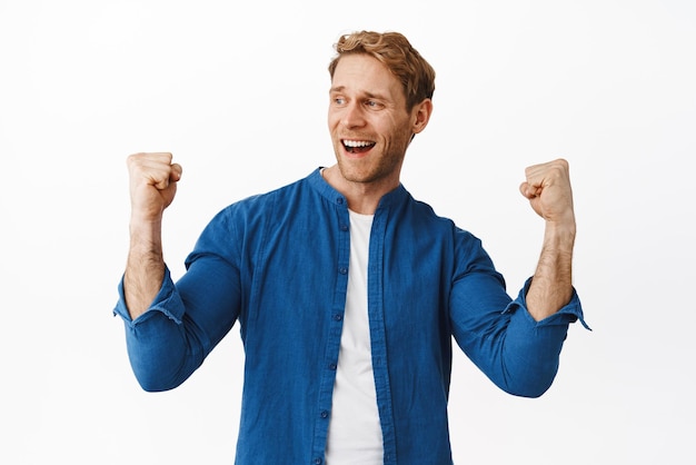 Free photo successful handsome man with red hair fist pump smiling and say yes looking left at winning prize achieve goal celebrate victory triumphing against white background