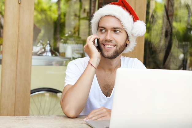 Successful and happy young man in red hat with white fur spending Christmas holidays in warm country, having phone conversation with his friend while using free wi-fi on generic laptop computer