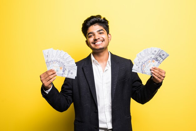 Successful Indian entrepreneur with dollar banknotes in hand classical suit looking at camera with toothy smile while standing against yellow wall