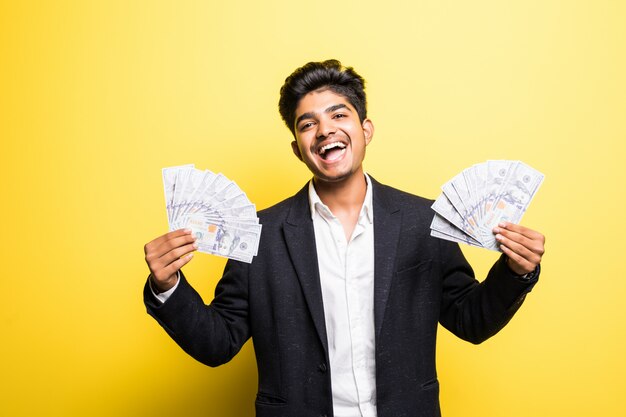Успешный индийский предприниматель с долларовыми банкнотами в классическом костюме руки смотрит на камеру с зубастой улыбкой, стоя у желтой стены