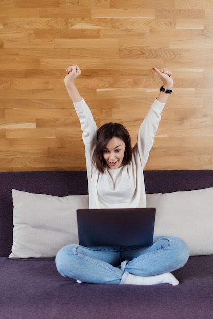 Foto gratuita la riuscita signora sta lavorando al computer portatile che si siede sul letto scuro davanti alla parete di legno a casa