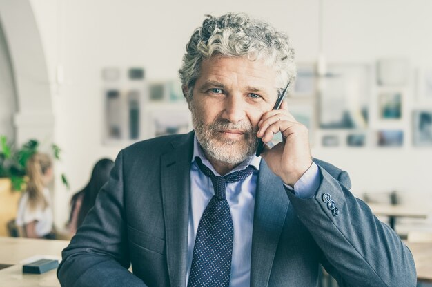 Успешный зрелый бизнесмен разговаривает по мобильному телефону, стоя в коворкинге, опираясь на стол