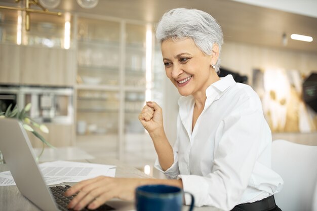 Successful mature female writer sitting at desk with portable computer, papers and cup of coffee, having happy facial expression because she managed to finish work in time