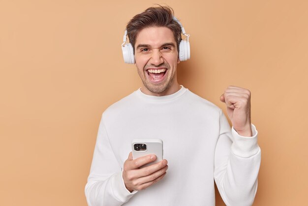 黒髪の成功したポジティブな男は歯を食いしばって良いニュースを喜ぶカジュアルな白いジャンパーはスマートフォンを使用してオンラインゲームをプレイし、ベージュの壁に隔離されたワイヤレスヘッドフォンを使用します。