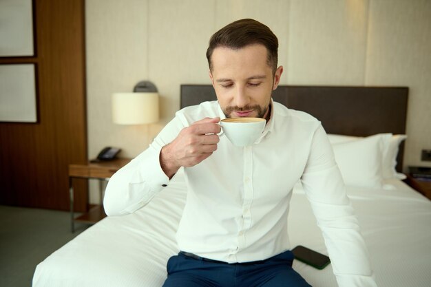 出張中にホテルのアパートでコーヒーを飲むビジネスカジュアルな服装で成功した中年の白人ビジネスマン。一日の始まりにぴったりの淹れたてのコーヒー