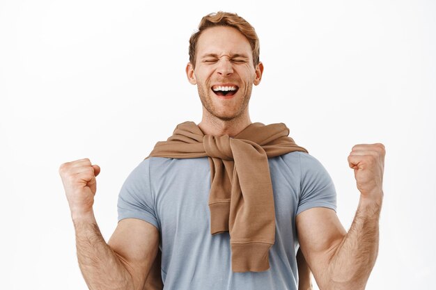 Successful redhead man winning, celebrating victory, shouting from joy and clench fists, make fist pump gesture to celebrate triumph, say yeah yes, win prize, standing over white background